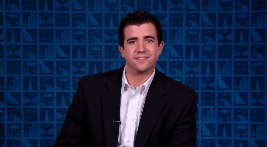 A smiling man with glasses, wearing a blue shirt, stands in front of a colorful light display.