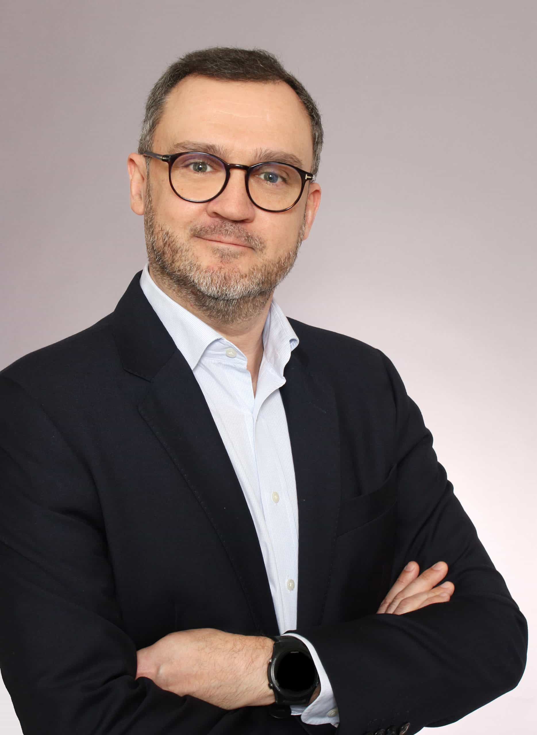 A man with short hair and glasses smiles confidently while standing in a modern office environment.