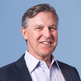 A man with glasses and a beard smiles confidently, wearing a blazer and collared shirt, against a neutral background.