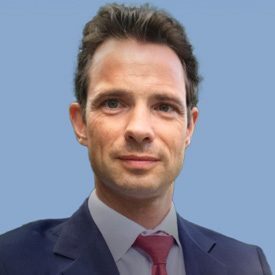 A man with glasses and a beard smiles confidently in a professional setting, wearing a dark blazer and white shirt.