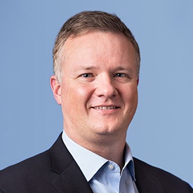 A man with glasses and a beard smiles confidently in a professional setting, wearing a dark suit and tie.
